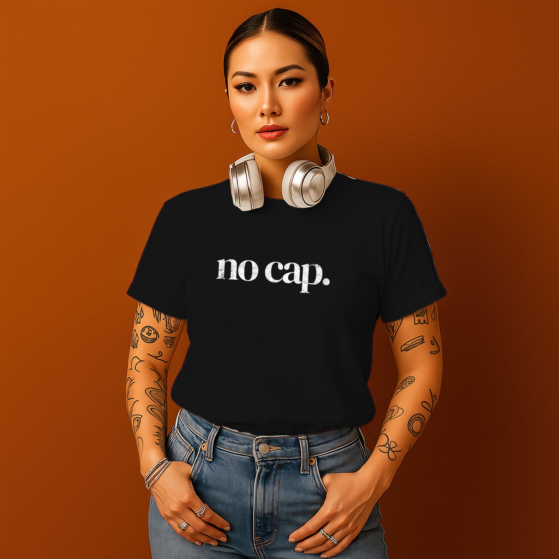 A woman with tattoos on her arms and a black t-shirt with the text "no cap" stands against an orange background, wearing headphones around her neck.