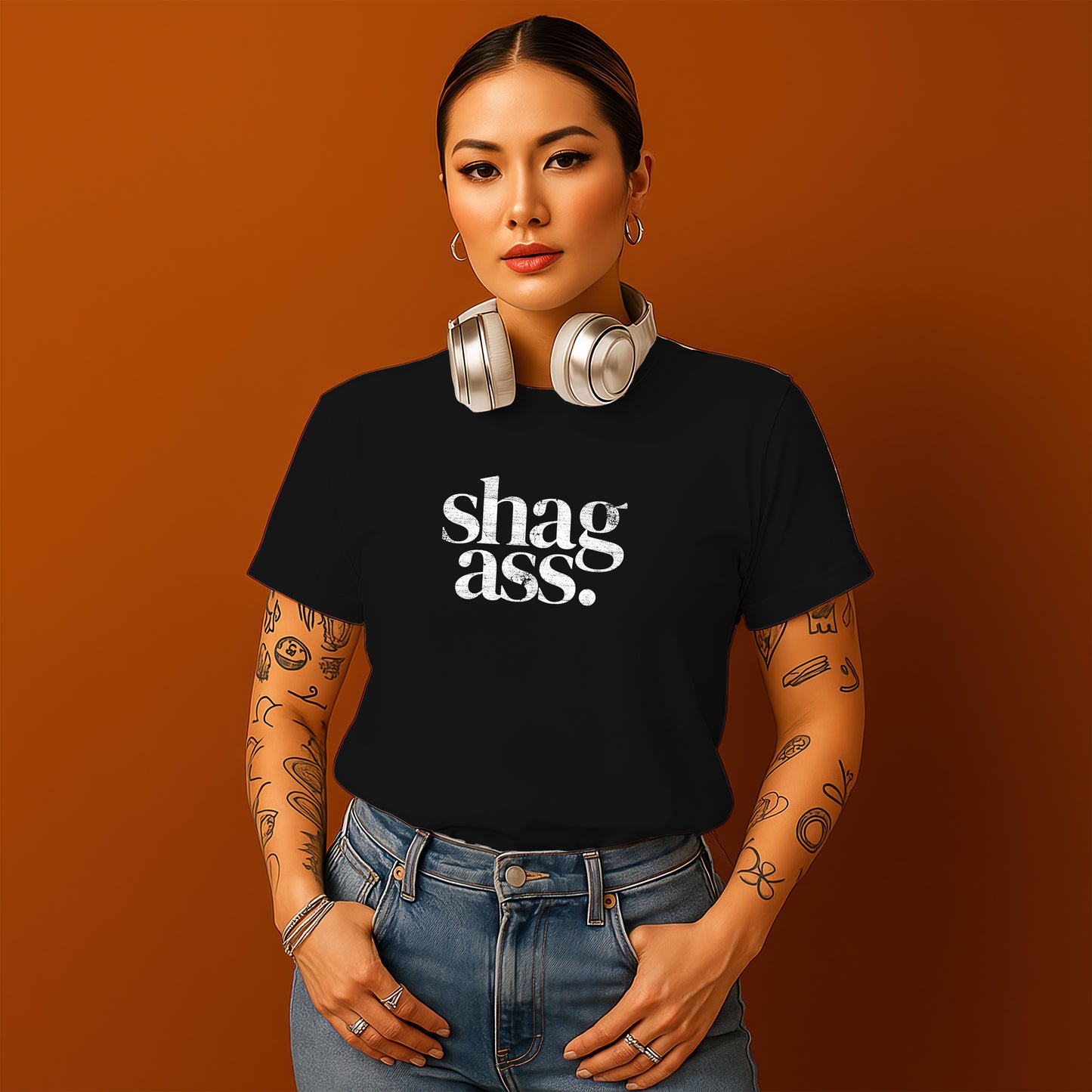 A woman with tattoos on her arms and a black t-shirt with the text "shag ass" stands against an orange background, wearing headphones and looking directly at the camera.