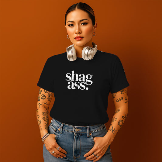 A woman with tattoos on her arms and a black t-shirt with the text "shag ass" stands against an orange background, wearing headphones and looking directly at the camera.