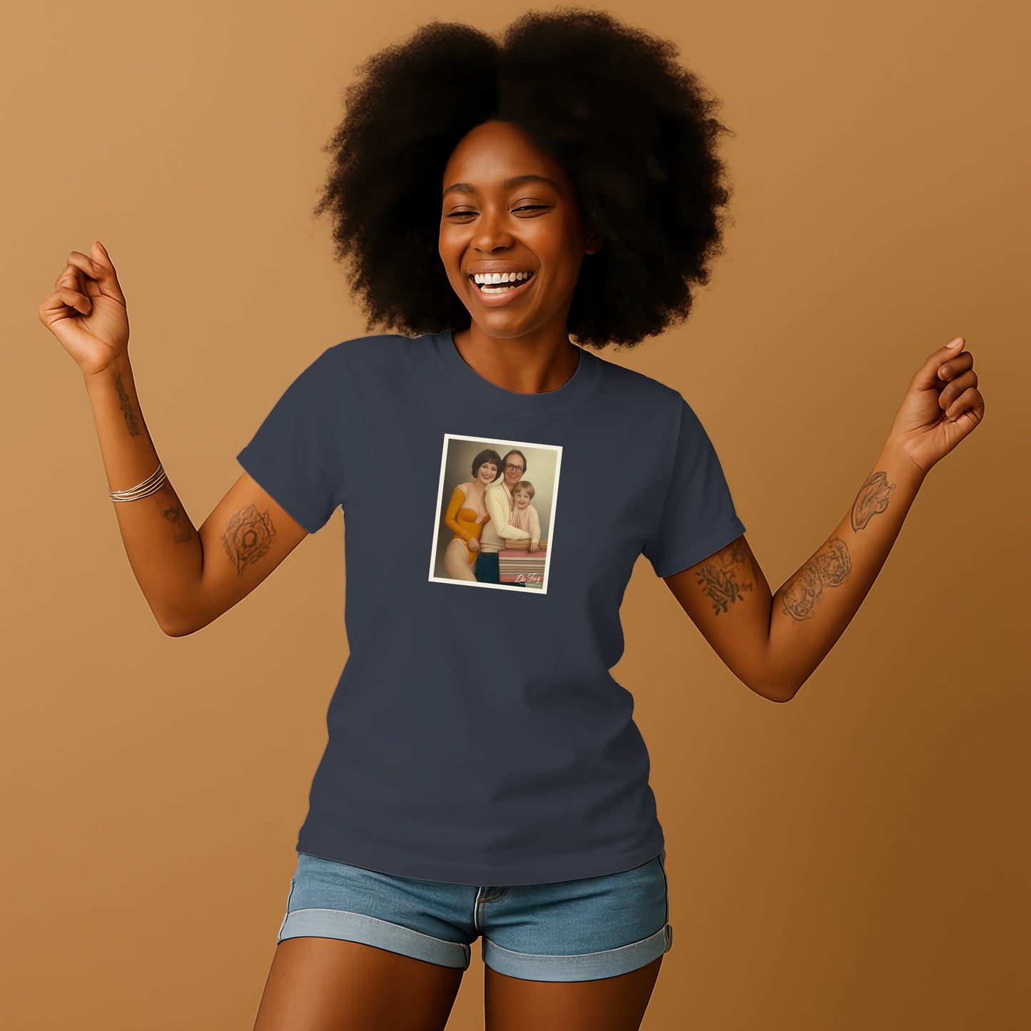 A smiling woman with curly hair wearing a blue t-shirt with a framed photo of two people on it, standing against a beige background.