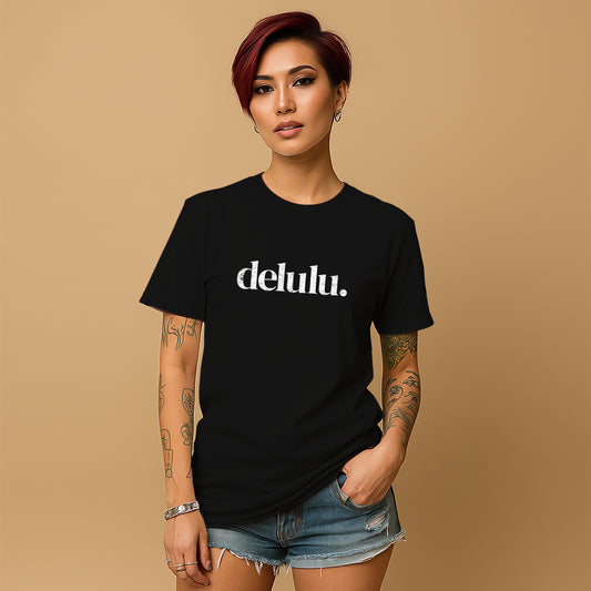 A young woman with short red hair wearing a black t-shirt with the word "delulu" printed on it, standing against a beige background.
